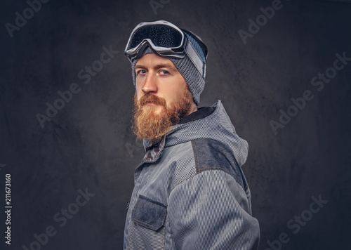 Portrait of a redhead snowboarder with a full beard in a winter hat and protective glasses dressed in a snowboarding coat posing at a studio, looking away. Isolated on dark textured background.