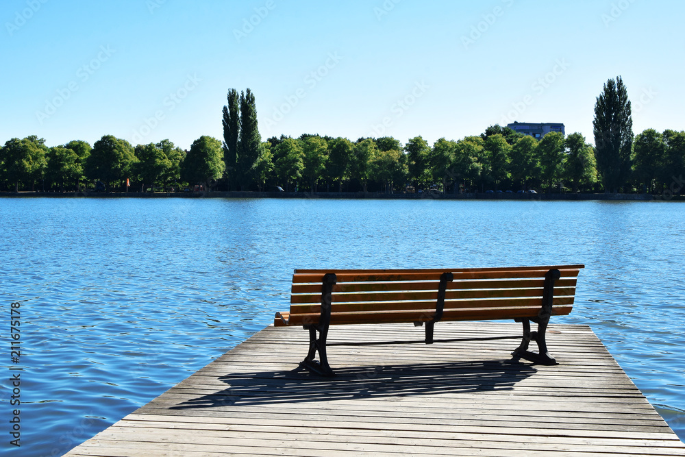 Empty bench on wooden deck with view on lake and green city park across the lake with copy space for text on shiny water.