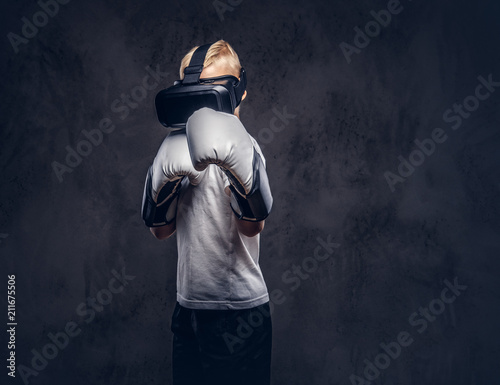 Schoolboy boxer with blonde hair dressed in a white t-shirt wearing visual reality glasses and boxing gloves, workout in a studio. Isolated on dark textured background.