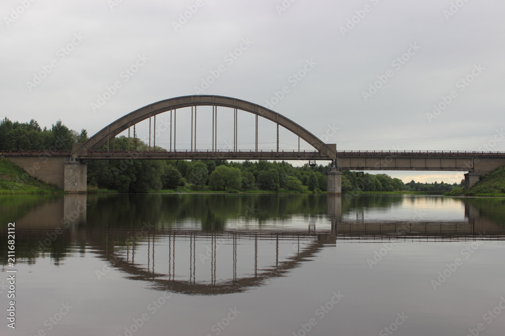 Classic old arched railway bridge over the river - view from the water on a summer day against the trees on the shore and gray cloudy sky