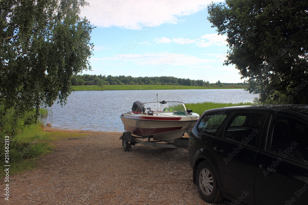 Outdoor activities - descent of a white-red motor plastic boat on a trailer with a car on the water on a sandy slipway on the background of the river and the shore with green trees
