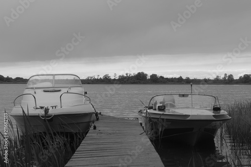 Two white old motor boats with cabin and open – moored at the jetty with the reeds in the evening on the background of the river and cloudy sky