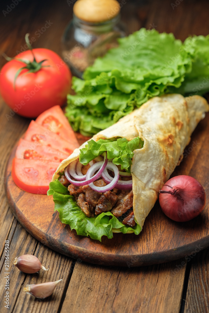 Doner kebab is lying on the cutting board. Shawarma with meat, onions, salad lies on a dark old wooden table. Doner kebab is lying on the cutting board. Shawarma with meat, onions, salad lies on a dark old wooden table.