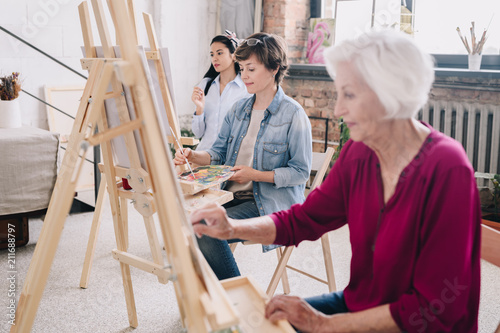 Portrait of art students sitting in row and painting at easels in art studio, focus on smiling  adult  woman enjoying work in center, copy space