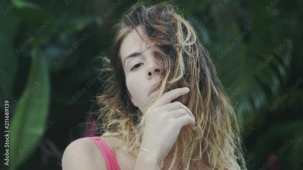 A close-up slow-motion picture of a girl with short, sunburned curly hair, covers them with a part of her face, sits amidst the tropical greenery, closes her eyes with pleasure. A close-up slow-motion picture of a girl with short, sunburned curly hair, covers them with a part of her face, sits amidst the tropical greenery, closes her eyes with pleasure.