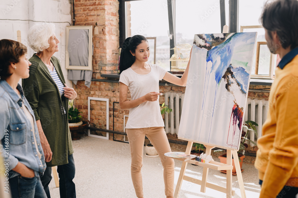 Fototapeta premium Portrait of pretty young woman presenting modern watercolor painting to audience standing in art-studio or gallery