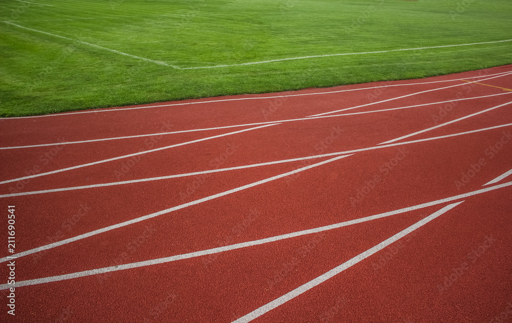Fototapeta premium red running road track with white marking lines near green football field and empty space for copy or text