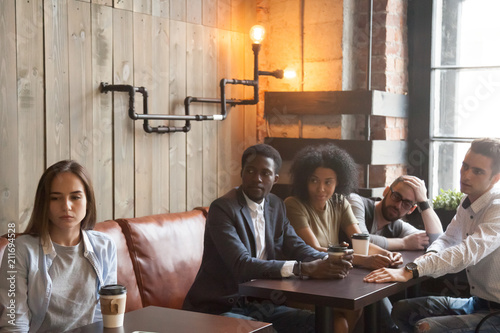 Upset millennial girl ignoring multiracial people trying to cheer her up, sitting at next table in coffeeshop, frustrated offended woman not talking with friends making peace with her in cafe