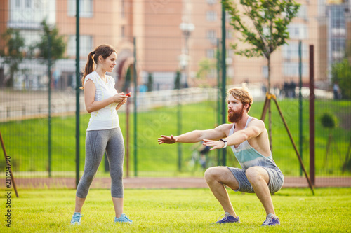 Fototapeta Naklejka Na Ścianę i Meble -  Team work in sports. A female trainer is training a young handsome man with a beard in a park on green grass, a lawn at sunset. A girl holds a phone in her hand and uses a stopwatch
