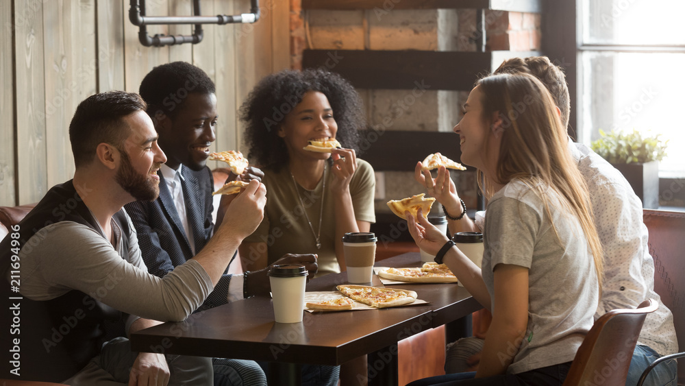 Smiling multiracial friends eating pizza and drinking coffee, laughing and having fun in restaurant, diverse millennial colleagues enjoying lunch during work break sitting at coffee table in loft cafe - obrazy, fototapety, plakaty 