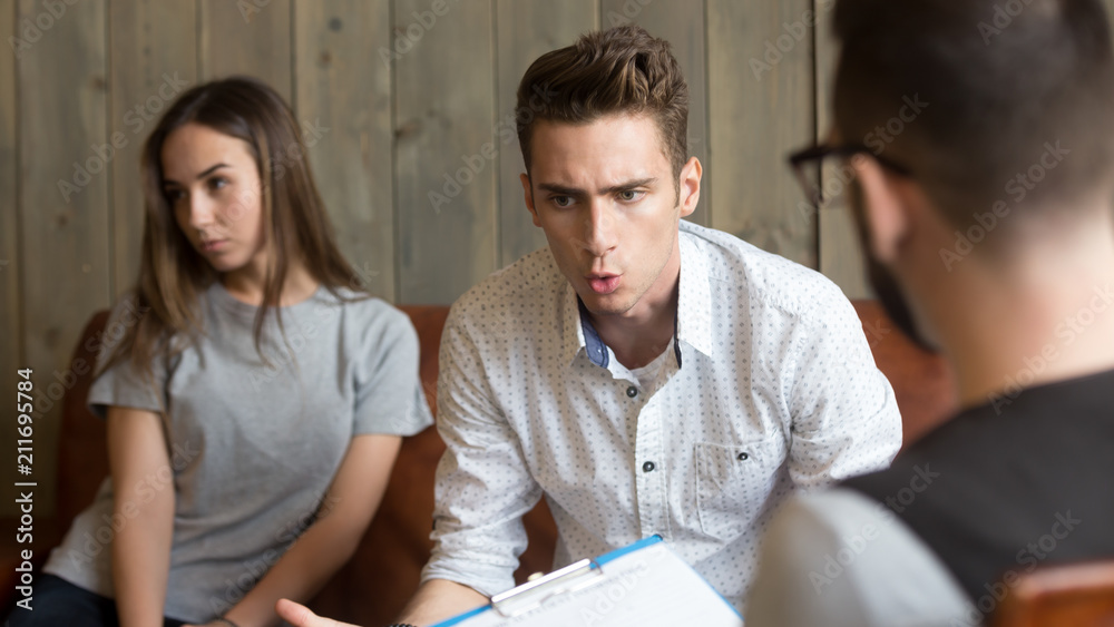 Mad Caucasian husband expressing complaints talking to psychologist during therapy session with wife, millennial couple undergoing family treatment at relationship counsellor office to save marriage Mad Caucasian husband expressing complaints talking to psychologist during therapy session with wife, millennial couple undergoing family treatment at relationship counsellor office to save marriage