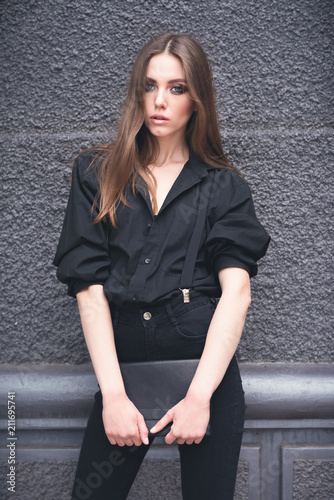 Beautiful girl with a black clutch in her hand. Woman in dark clothes on a dark background in the street. Toned image.