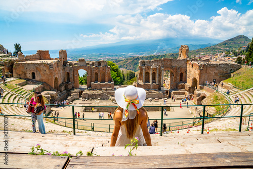 Jule 04, 2018. Taormina, Italy. Beautiful girl in a long white dress and white summer hat sitting at the caldron of the ancient Greek Theater of Taormina in Italy. photo