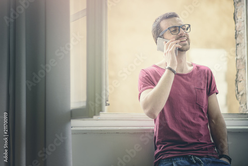 Happy young man talking on his cell phone, standing next to his big home window