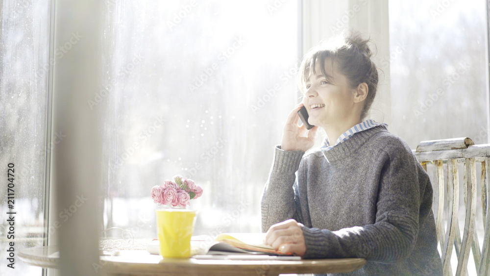 A young smiling laughing cute brunette girl dressed in a blue pullover is sitting near the window having a call A young smiling laughing cute brunette girl dressed in a blue pullover is sitting near the window having a call