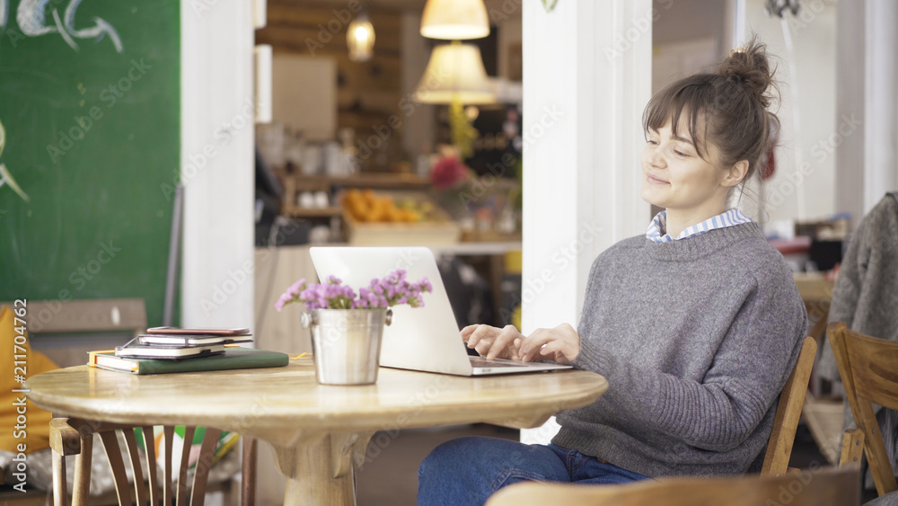 A young smiling cute happy brunette girl dressed in a grey pullover is working with a laptop in a cafe A young smiling cute happy brunette girl dressed in a grey pullover is working with a laptop in a cafe