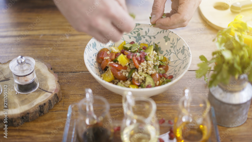 Male chief cook's hands adding parsley to a salad bowl of cucumbers, tomatoes and paprika and seeds