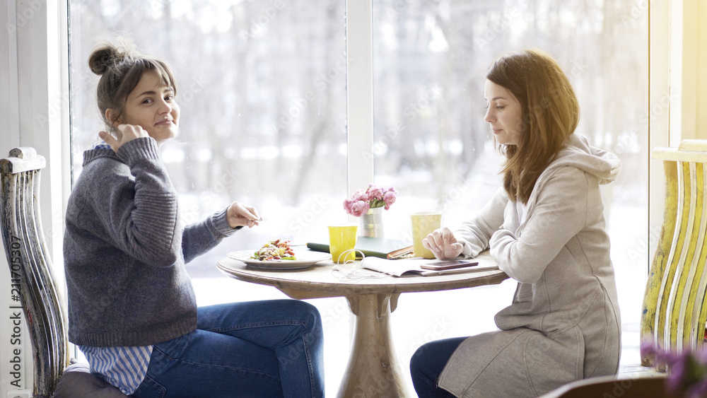 Two female friends having a conversation in a cafe drinking coffee eating salad looking at the camera Two female friends having a conversation in a cafe drinking coffee eating salad looking at the camera