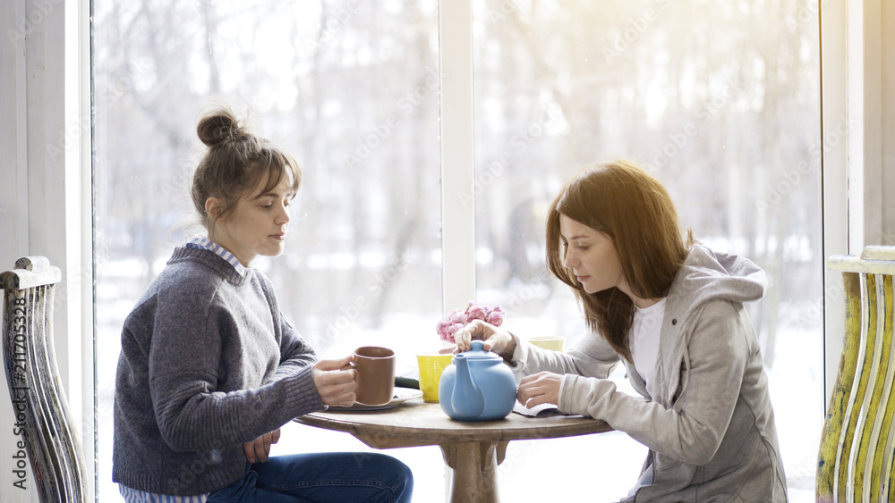 Two female friends adult girls sitting in front of each other drinking tea in a cafe holding closing a blue tea pot Two female friends adult girls sitting in front of each other drinking tea in a cafe holding closing a blue tea pot