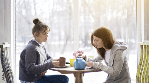 Two female friends adult girls sitting in front of each other drinking tea in a cafe holding closing a blue tea pot