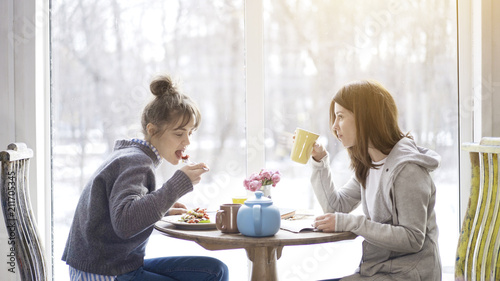 Two young adult female friends eating salad drinking tea sitting near a big window indoors in a cafe
