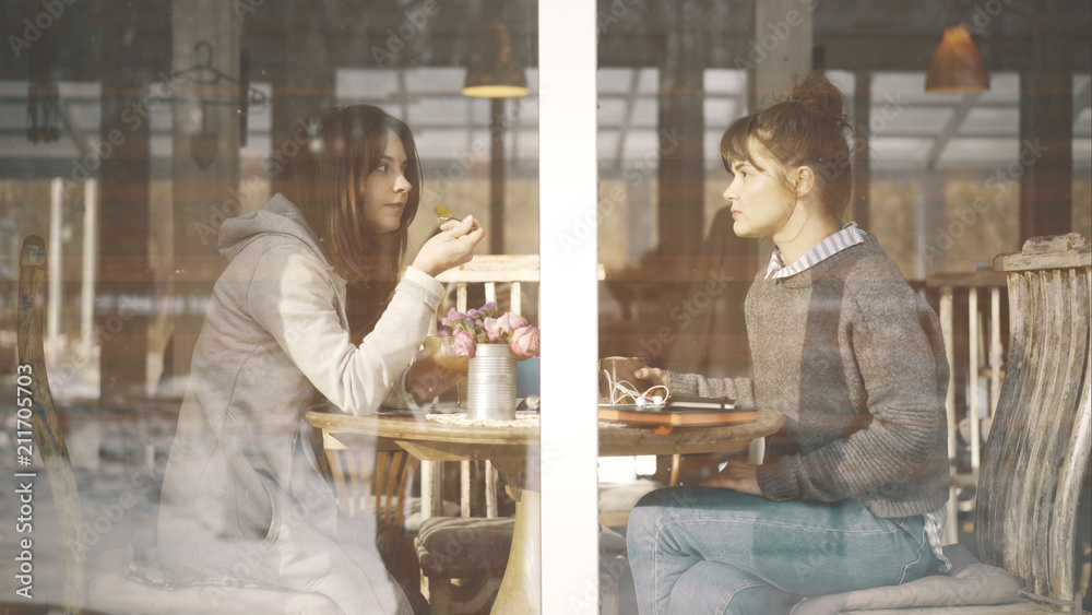 Two casual cute female friends smiling having a serious talk on a sunny day met in a cafe to talk and eat dessert and drink tea