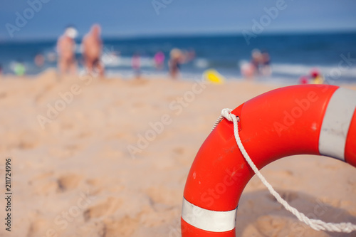 Orange lifebuoy in foreground. Blue clear sky, sea and people in background. Bright sunny day. Holidays at beach. Beautiful seascape. Equipment for rescue. Service for lifesaving.