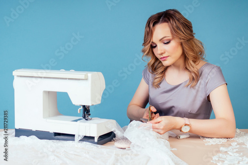 attractive woman seamstress tailor ( dressmaker) holds scissors and cuts fabric on table with sewing machine on a blue background in the studio. photo