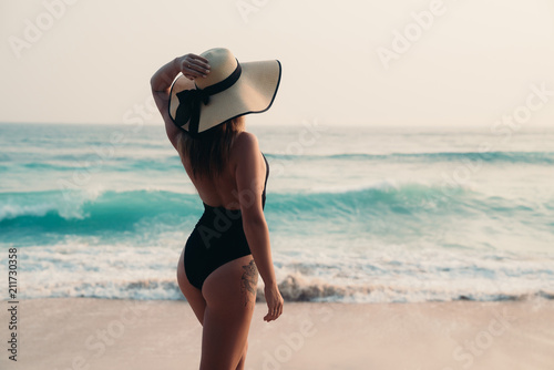 View from the back of a slender young girl in a fashionable bathing suit, wears a stylish beach with wide fields, holds her hand from the wind from the sea, admires the waves on the beach