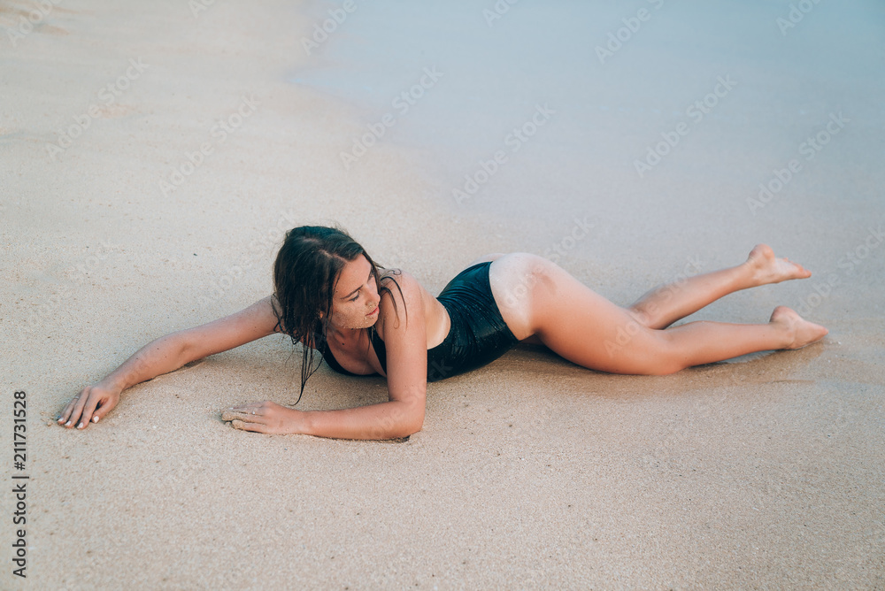 Fototapeta premium A sexy young girl with a smooth even skin in a fashionable black swimsuit lies on wet sand. The concept of leisure, travel, people.