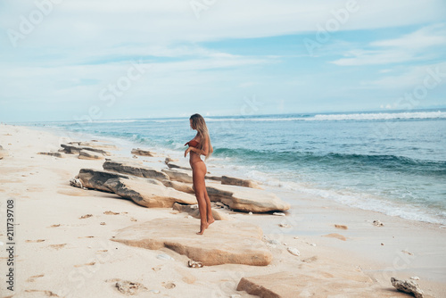A sexy model in a swimsuit stands in profile and admires the rocks on a deserted white beach. A girl with an even tan and a beautiful body came to relax.