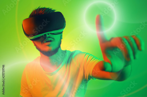 Attractive bearded man trying VR headset and poniting in lightning circle. Young man exploring another world with virtual reality goggles on the colorful background. photo