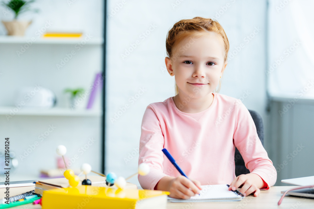beautiful redhead schoolgirl smiling at camera while writing in notebook at desk beautiful redhead schoolgirl smiling at camera while writing in notebook at desk