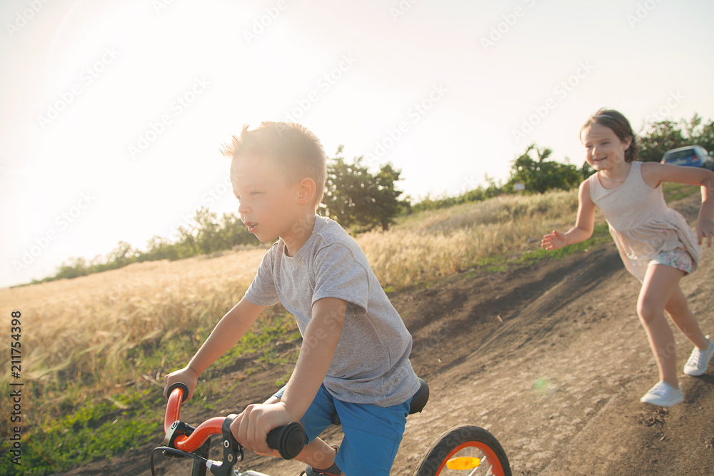 Boy and girl in the field. The boy is riding a bicycle, and the girl is running alongside. A cheerful, happy childhood in the village. - obrazy, fototapety, plakaty 
