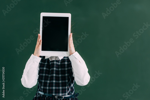 schoolchild hiding face with digital tablet while standing near chalkboard