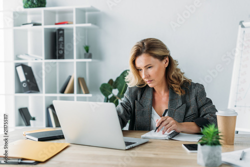 beautiful businesswoman using laptop and writing something to notebook in office