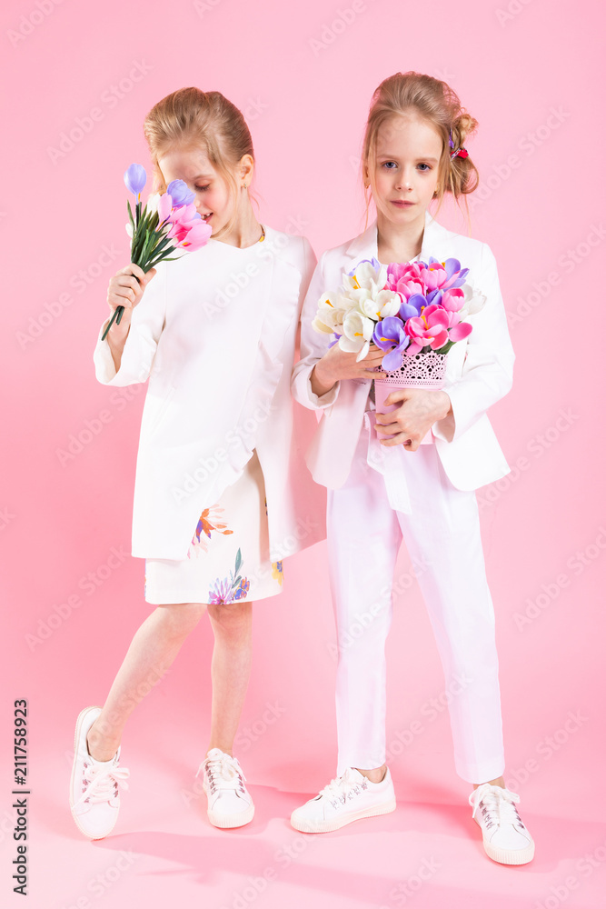 Twins girls in light clothes with bouquets of flowers stand on a pink background. Twins girls in light clothes with bouquets of flowers stand on a pink background.