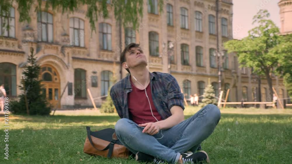 Young handsome man is sitting on grass in park, listening music with earphones, relax concept, dreaming concept, building on background Young handsome man is sitting on grass in park, listening music with earphones, relax concept, dreaming concept, building on background