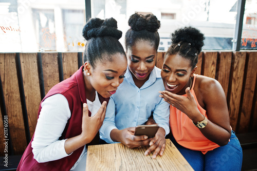 Three african american girls sitting on the table of caffe and looking on mobile phone.