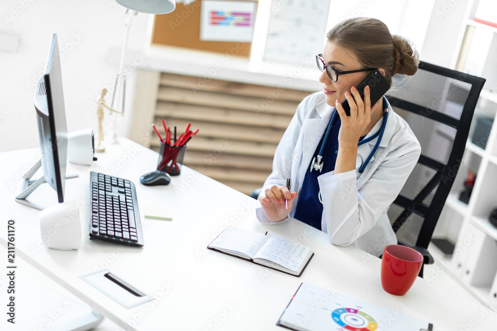 Φωτογραφία A young girl in a white robe is sitting at the desk in the office, talking on the phone and holding a pen in her hand