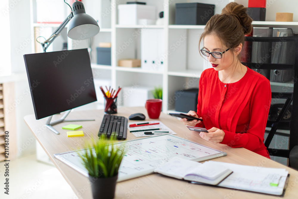 A young girl is sitting at the desk in the office, holding a bank card and phone in her hand. A young girl is sitting at the desk in the office, holding a bank card and phone in her hand.