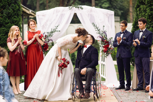 Bride and groom on the wheelchair kiss before wedding altar after the ceremony