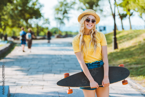 portrait of smiling blond woman in sunglasses and hat with longboard in hands on street photo