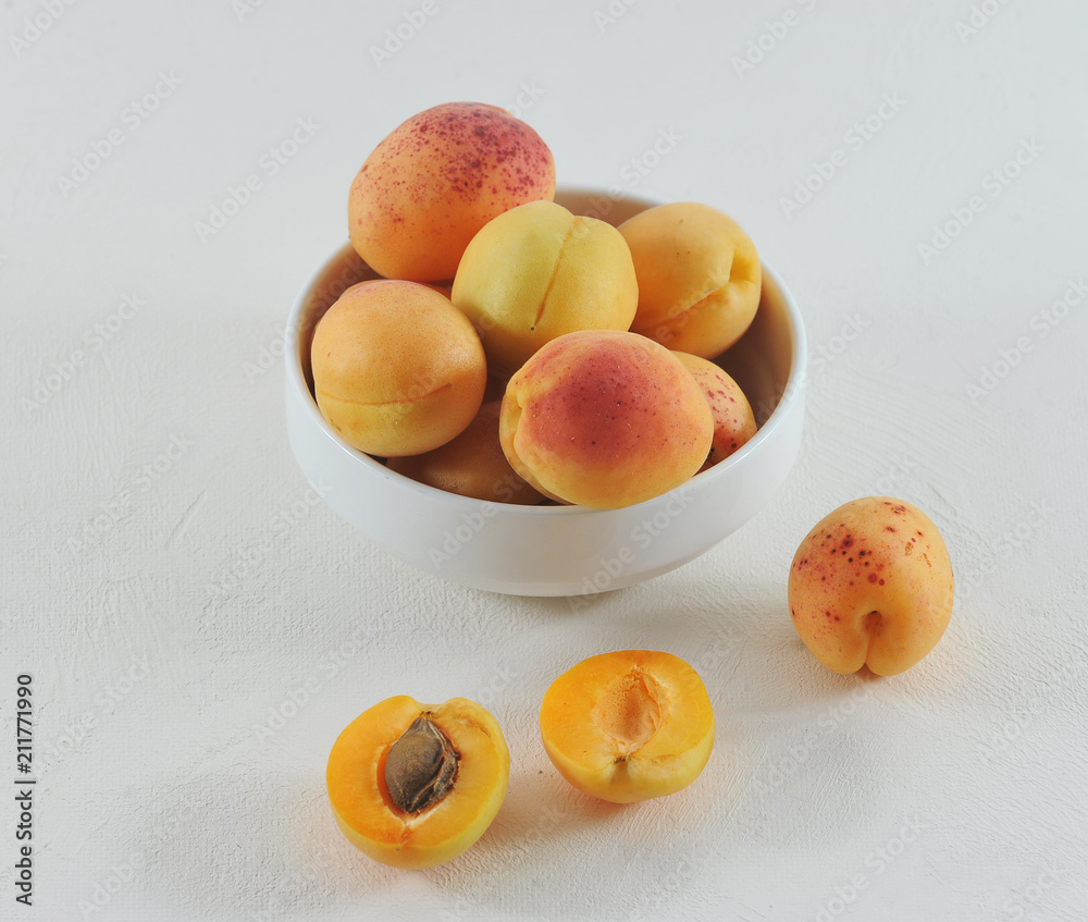 Ripe apricots in a white porcelain bowl. Several apricot fruits lie next to the bowl. One apricot is broken in half. Close-up. White background. Ripe apricots in a white porcelain bowl. Several apricot fruits lie next to the bowl. One apricot is broken in half. Close-up. White background.