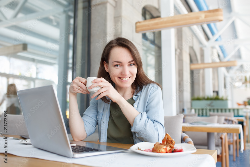 Portrait of beautiful young woman accountant or manager of large company sitting at table in cafe during lunch with mug of coffee and social networking using a laptop and wifi Portrait of beautiful young woman accountant or manager of large company sitting at table in cafe during lunch with mug of coffee and social networking using a laptop and wifi