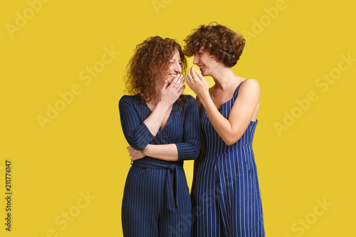 Handsome young caucasian student in dark blue overalls standing at yellow background, whispering something into ear of stylish ginger girl, sharing latest gossip. Woman looking in happy and smiling