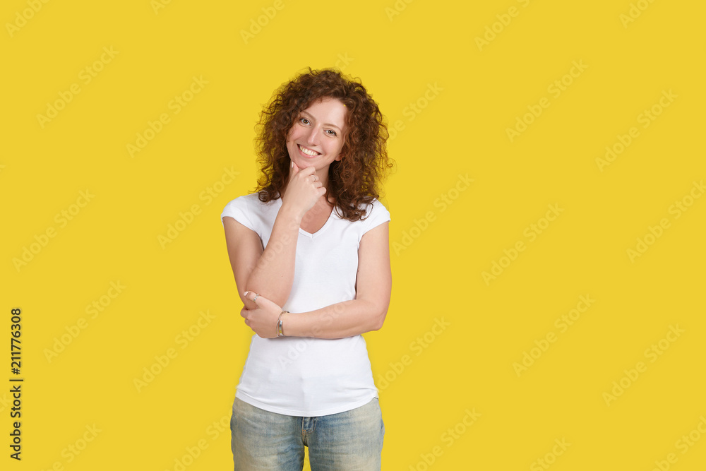 Friendly happy caucasian American teenager wearing casual white T-shirt holding her hand on chin having joyful and cheerful look, smiles gently, stands on yellow wall. People, positive emotions Friendly happy caucasian American teenager wearing casual white T-shirt holding her hand on chin having joyful and cheerful look, smiles gently, stands on yellow wall. People, positive emotions