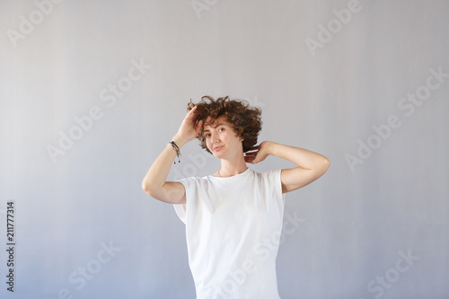 Young wonderful girl keeps herself for curly head, rejoices in her youth, has great good mood. Copy space, gray studio background. Happy joyful female student smiling