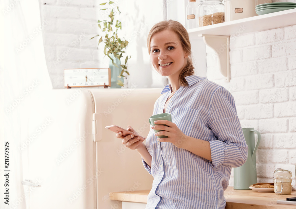 Young woman using smartphone leaning at kitchen table with coffee mug and organizer in a modern home. Smiling woman reading phone message. Brunette happy girl typing a text message. Young woman using smartphone leaning at kitchen table with coffee mug and organizer in a modern home. Smiling woman reading phone message. Brunette happy girl typing a text message.