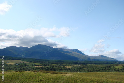 Panorama of the Scottish Forest at the foot of the mountain ranges against the background of a cloudy blue sky.